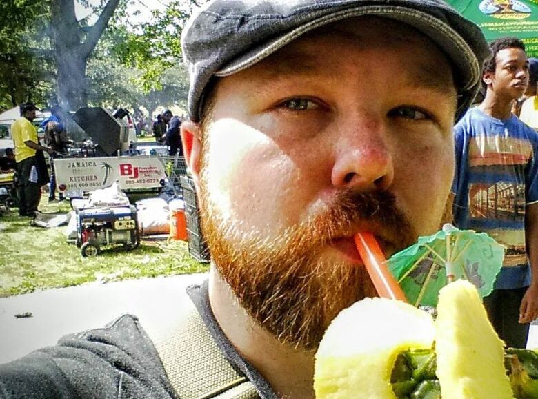 A bearded man with a hat takes a selfie of himself drinking from a pineapple