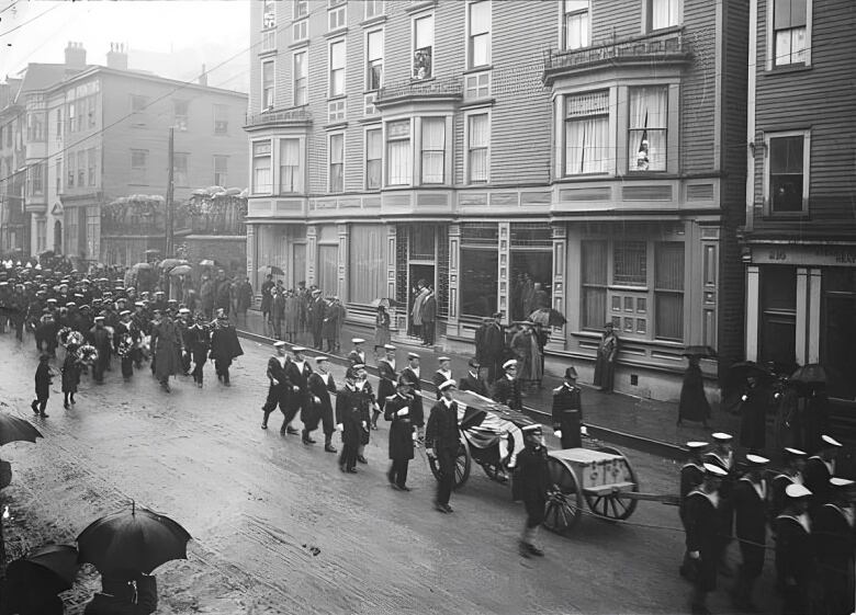 A black-and-white picture of mourners marching down a street, wheeling two caskets.