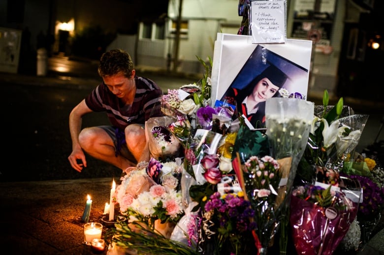 A man kneels beside a collection of flowers and candles laid on the ground, surrounding a graduation picture of a young woman.