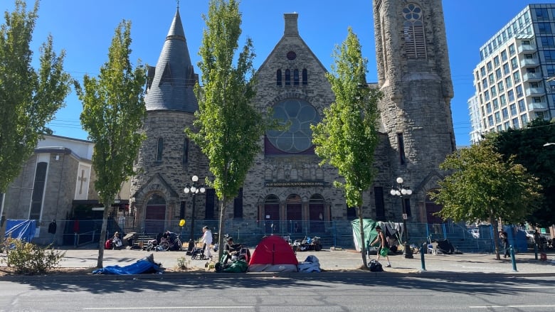 A temporary blue metal fence surrounds a large grey stone cathedral, with people camped out by the fence.