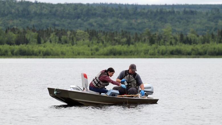 A man and woman wearing life jackets in a small boat with bottles and other equipment on a lake with green trees in the background.