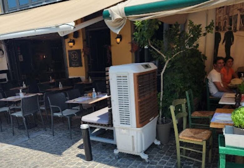 An outdoor air conditioner sits on a patio. People sit in a shaded part of the patio nearby.