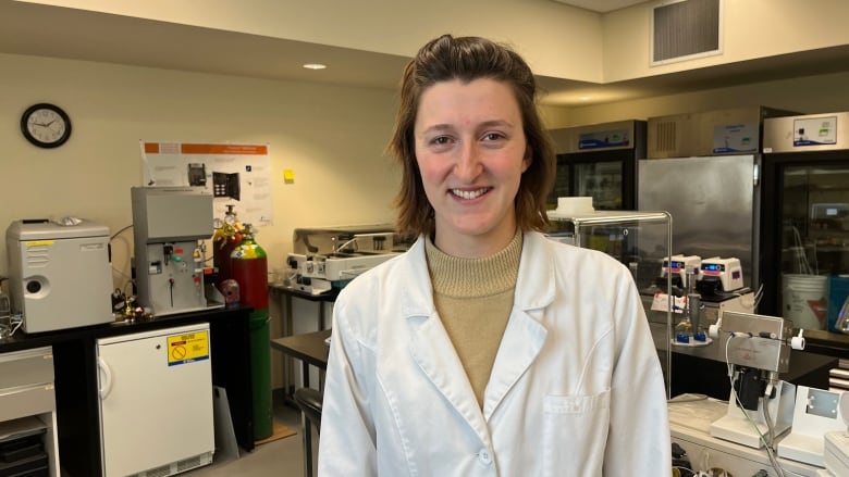 A young woman in a white lab coat stands inside a laboratory.