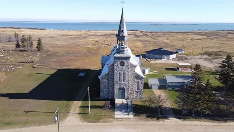 An aerial shot shows a stone church in an open field.