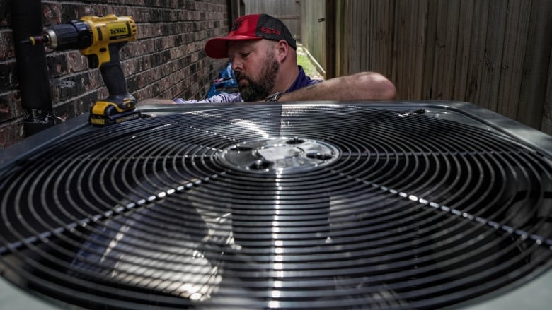 A man with a dark beard, wearing a red and black ball cap, fixes an outdoor air-conditioning unit.