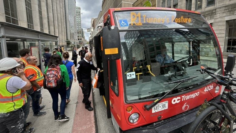 People board a red and white transit bus in a city's downtown in summer.