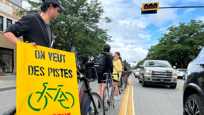 People stand in line with their bikes down a road. The person closest to the camera holds a sign saying 