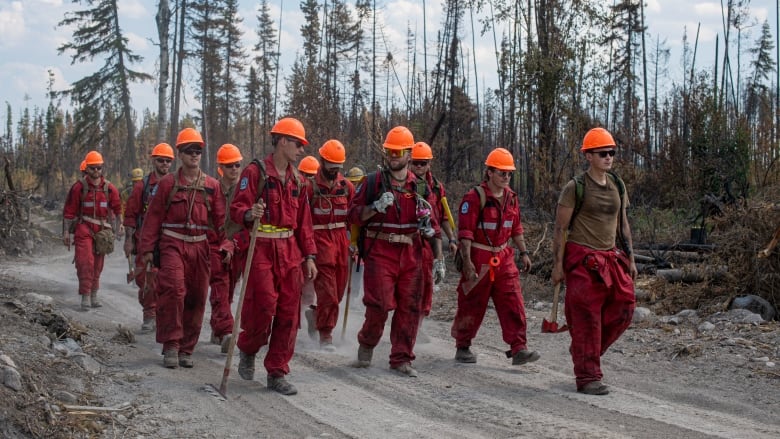 A line of men wearing red firefighting suits walk along a forest path.