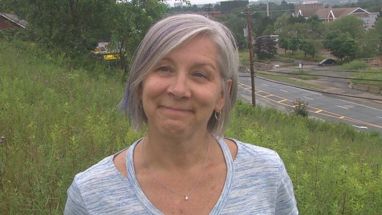 A woman in a blue and white shirt stands on a hill with a road in the background.