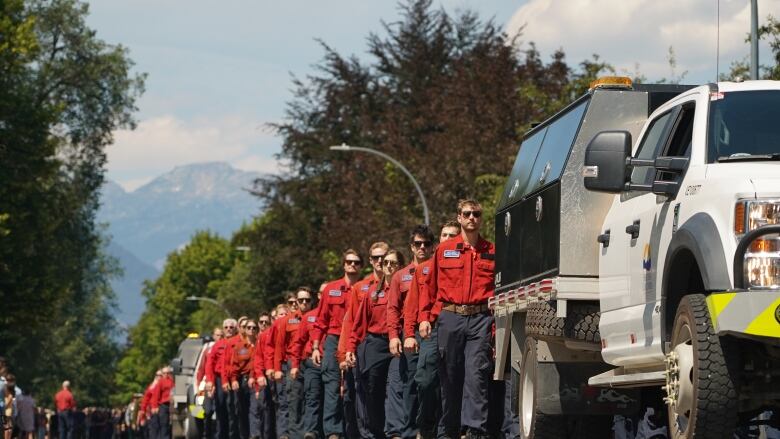 A line of firefighters in red shirts and blue pants marched behind a white truck with a black cab.