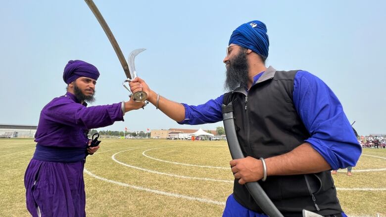 Two men wearing turbans hit swords together in a field.