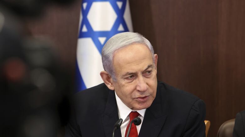 A person speaks into a microphone as the Israel flag is seen behind them.