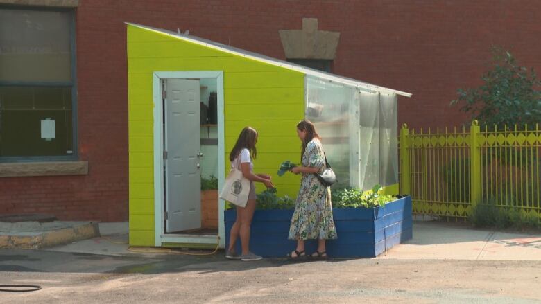 Two women stand in front of a green structure with plants in front. 