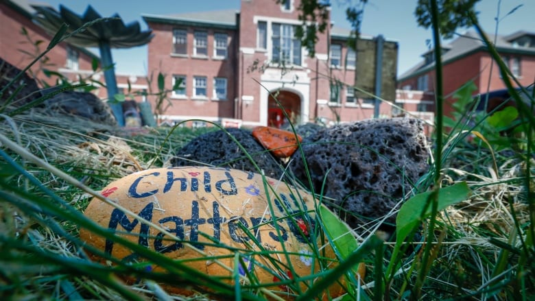 A hand-painted stone that says 'Every Child Matters' is seen amongst grass in the foreground, in front of a large red brick building.