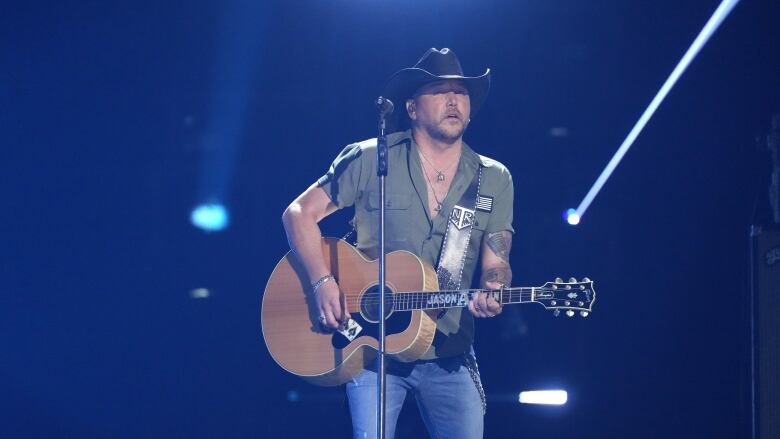 A man in a cowboy hate holds an acoustic guitar while onstage.