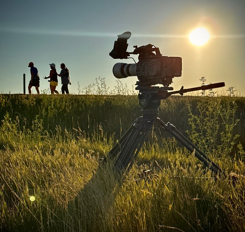 A group of three people in silhouette run on a grassy hill. A film camera on a tripod sits on the hill in the foreground.