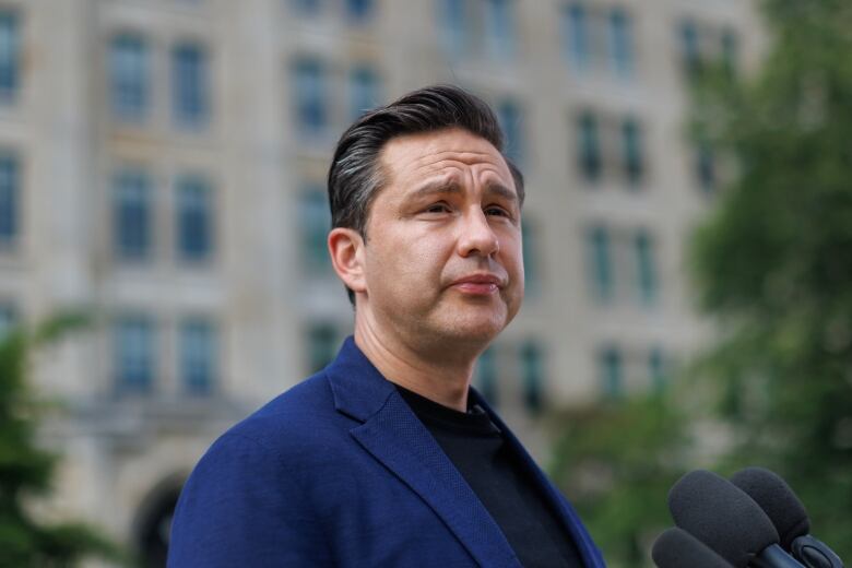 A man in a dark blue blazer and black t-shirt stands in a park with a blurry building int the background.