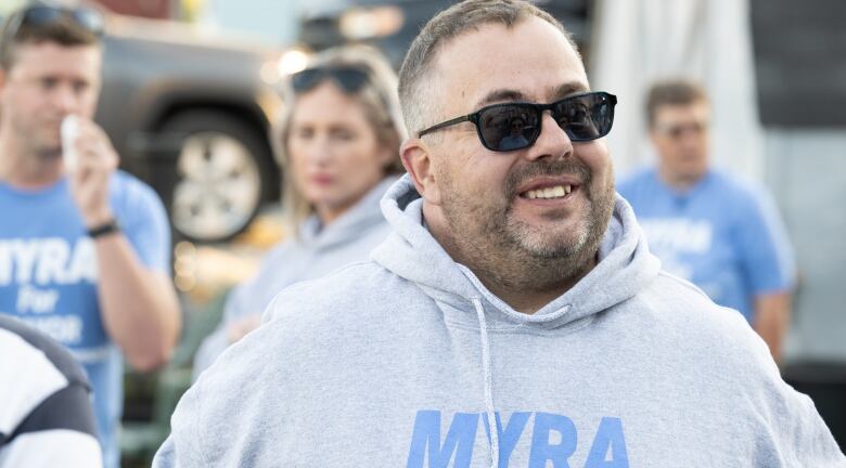 A white man in sunglasses and a grey hoodie sweatshirt smiles as other in blue campaign shirts are seen out of focus behind him.