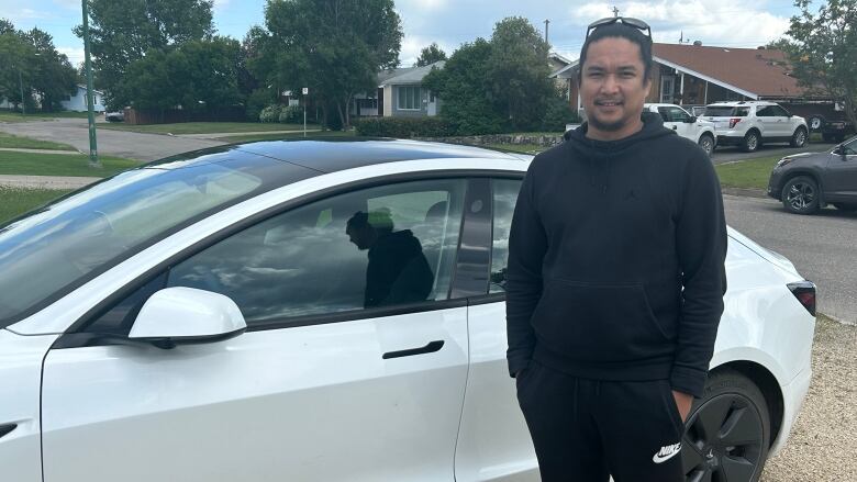 A man in a black sweatsuit standing in front of a white car. 