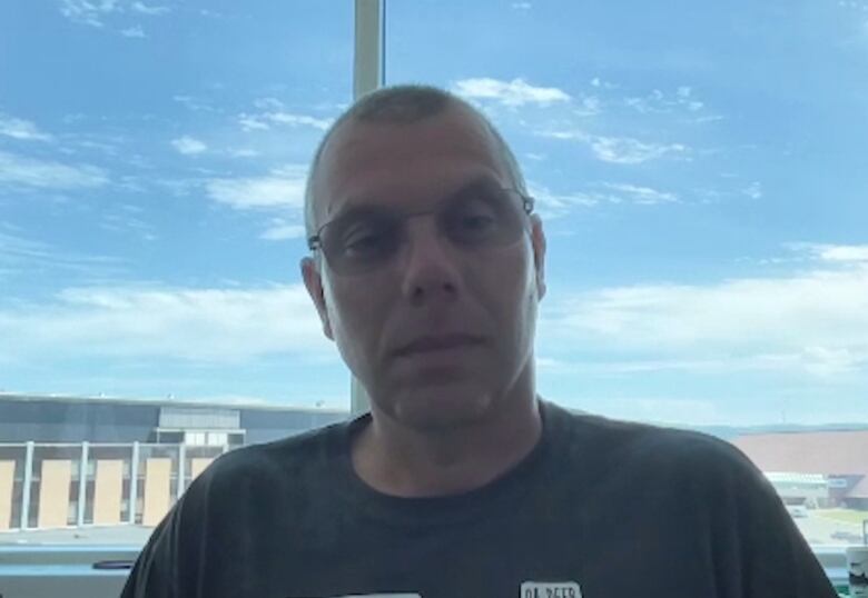 A bald man with glasses sits in front of a large window overlooking the Memorial University campus.