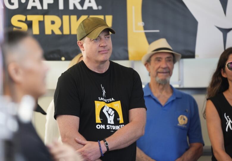 A man wearing a baseball cap and black shirt is shown in the foreground, while an older man wearing a fedora and blue shirt stands behind him.