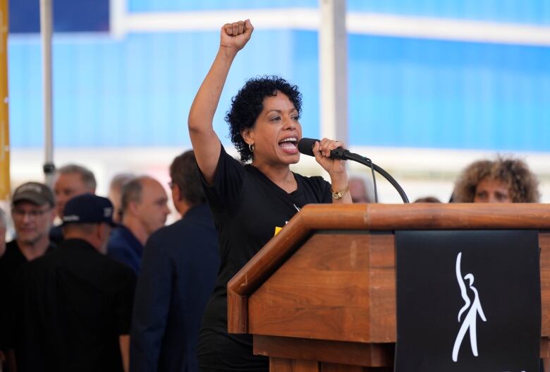 A woman with short curly hair stands at a podium and speaks into a microphone, raising a fist in the air.