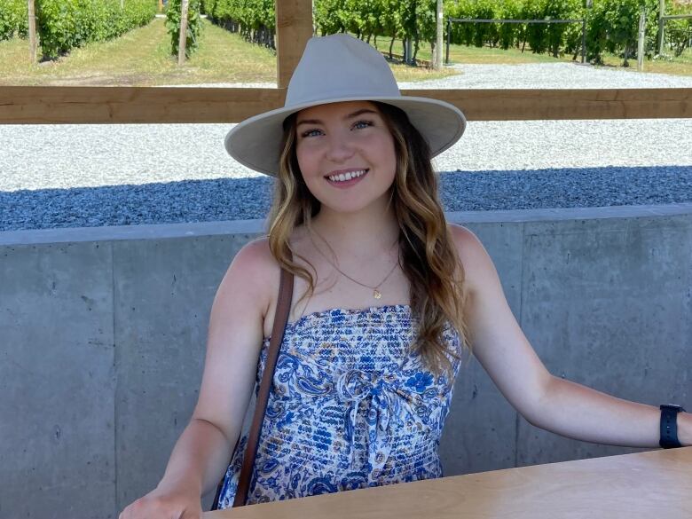 Emma Hamilton sits at an outdoor table and is wearing a blue and white strapless dress and a cream-coloured hat.