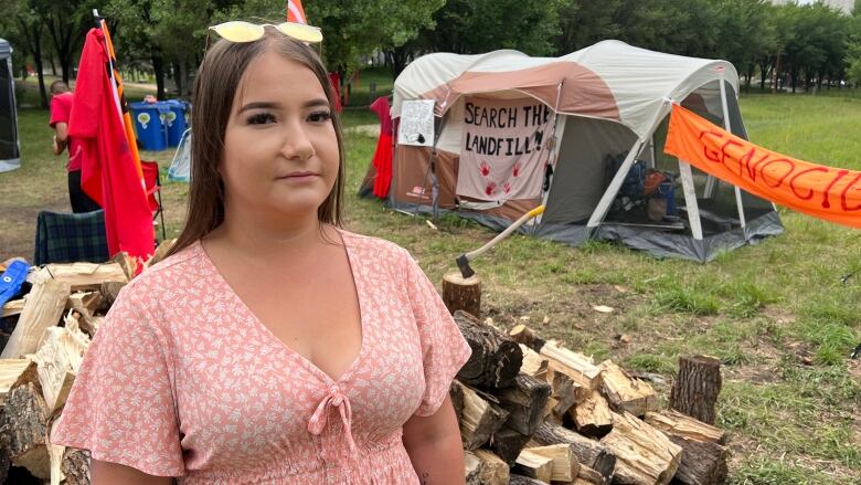 A woman stands in front of a pile of firewood and a tent with a sign that says 