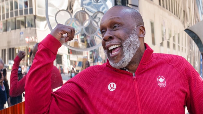 A man flexes and smiles in front of the Olympic rings.