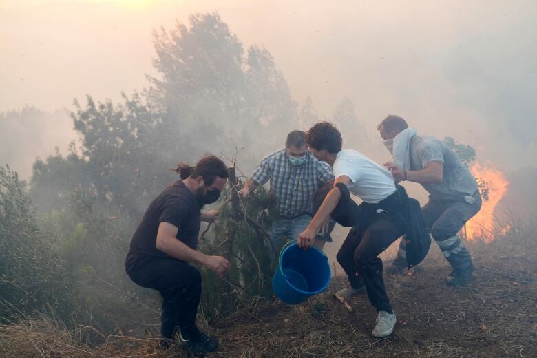 Four men crouch on a smoky hillside with flames nearby. One grips a large blue bucket in his left hand.