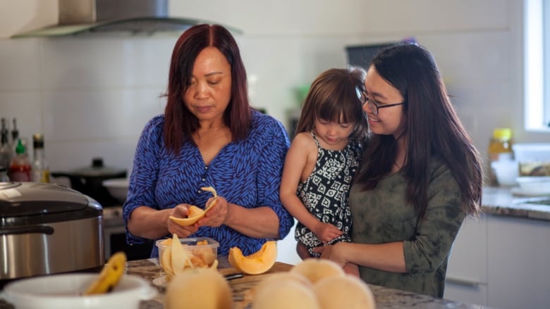 A woman peels fruit while her daughter and grand-daughter look on. 