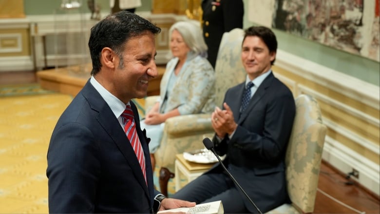 Minister of Justice and Attorney General of Canada Arif Virani wears a black suit and red tie. He is hold a book and is taking an oath of office. Prime Minister Justin Trudeau looks on wearing a dark suit and blue tie.