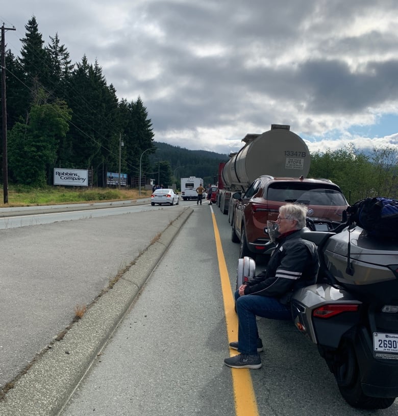 A man sits on a motorcyle without his helmet on waiting and staring with a long line of vehicles in front of him on a rural stretch of highway.