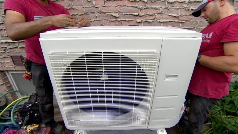 Two workers install a white heat pump outside in the backyard of a house.