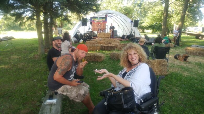 A tattooed person in a red baseball cap and camouflage shorts sits beside a person with long curly hair and a big smile. They appear to be holding cannabis paraphernalia.