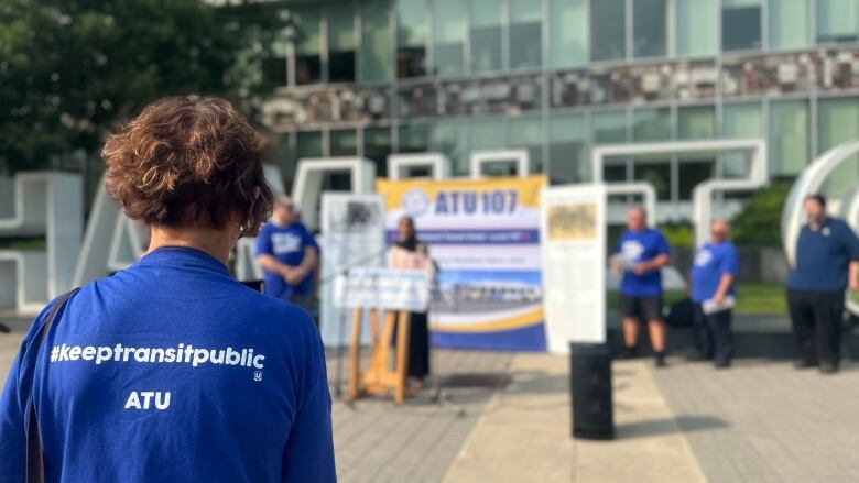 Woman speaking in front of Hamilton sign and other woman is taking her video in a #keeptransitpublic shirt.