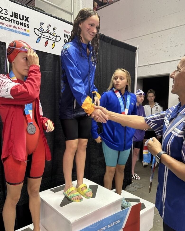 A young woman stands on a podium smiling while she shakes hands with a male official holding a gold medal he is about to present her with. 