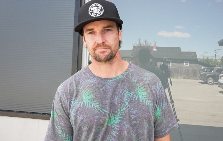 A man in a grey T-shirt and black cap, with brown facial hair, looks into the camera.