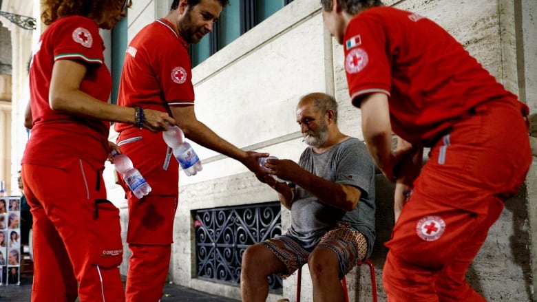 Three heath-care workers in red uniforms hand bottled water to a man seated outside a building. He is visibly sweating, his clothes damp. 