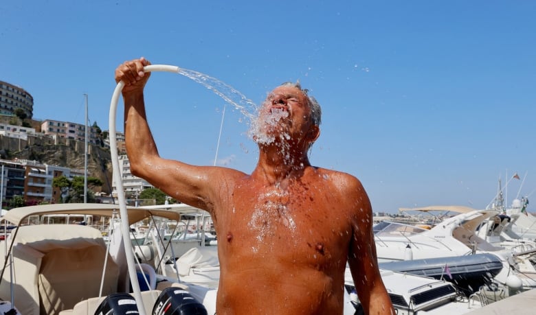 A shirtless man sprays his face and upper chest with water from a hose. Many boats and seaside buildings are in the background. 