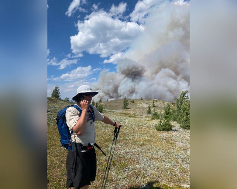 A man with hat, T-shirt and shorts is talking on the cellphone while standing on a hill, with a large plume of smoke in the background.