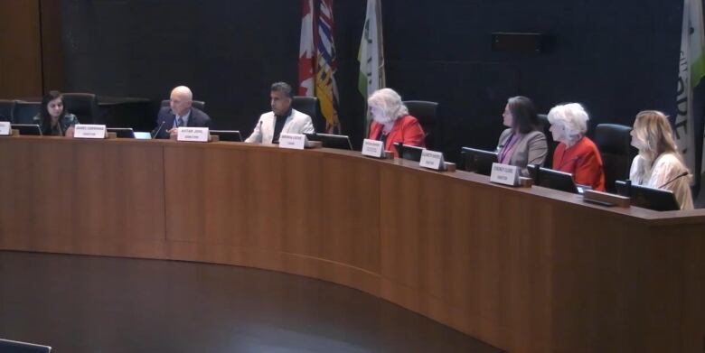 Seven people sit in an official council chamber in front of flags of Canada, B.C. and Surrey.