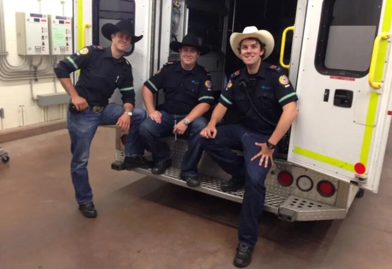 Three men pose for the camera at the back of an ambulance. They are wearing jeans and cowboy hats. 