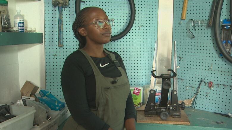 A woman wearing glasses and a black shirt with green overalls stands in front of bike repair supplies.