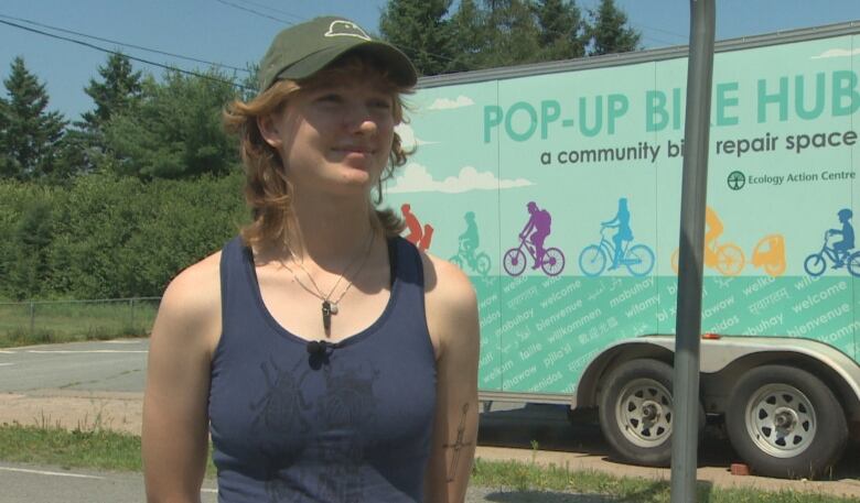 A woman wearing a green ballcap and a blue tank top poses for a photo next to the Pop-Up Bike Hub trailer.