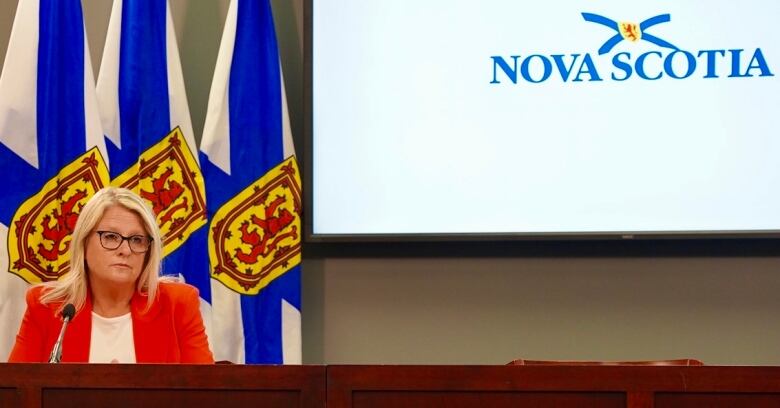 A woman with glasses sits at a podium with flags behind her.