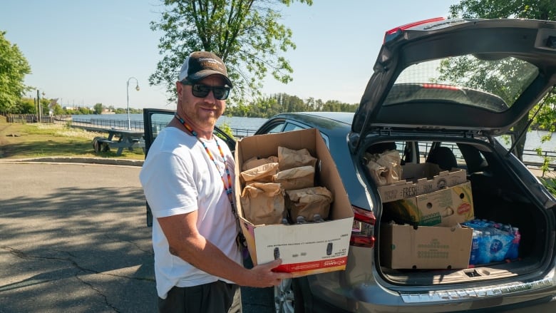 A man stands outside the open trunk of a car and holds a box of bagged food and water bottles.