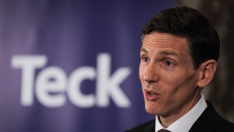 A middle-aged man with brown hair parted in the middle and sculpted cheekbones wears a black and white suit in front of a sign that says Teck.