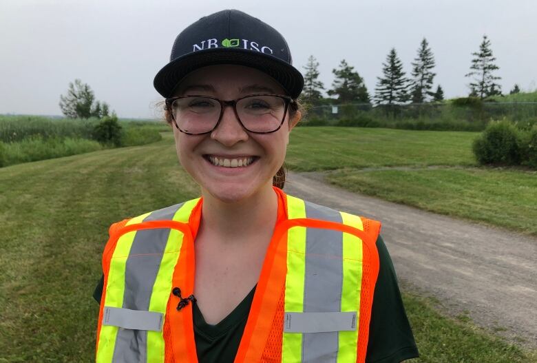 A smiling woman wearing an orange and yellow safety vest and a baseball cap that says NBISC.