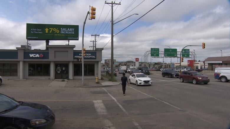 A billboard can be seen in a shopping plaza, with cars driving by on the road and a person is crossing the street in front of the billboard. 